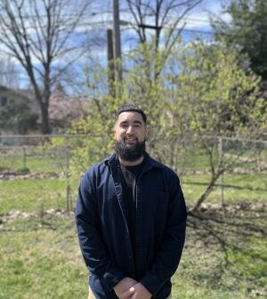 A man stands, hands clasped, outside in a nature space.