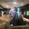 Main exhibit room for In Her Closet featuring drag costumes on grey mannequins. Text descriptions printed on multiple colored posters along black walls.